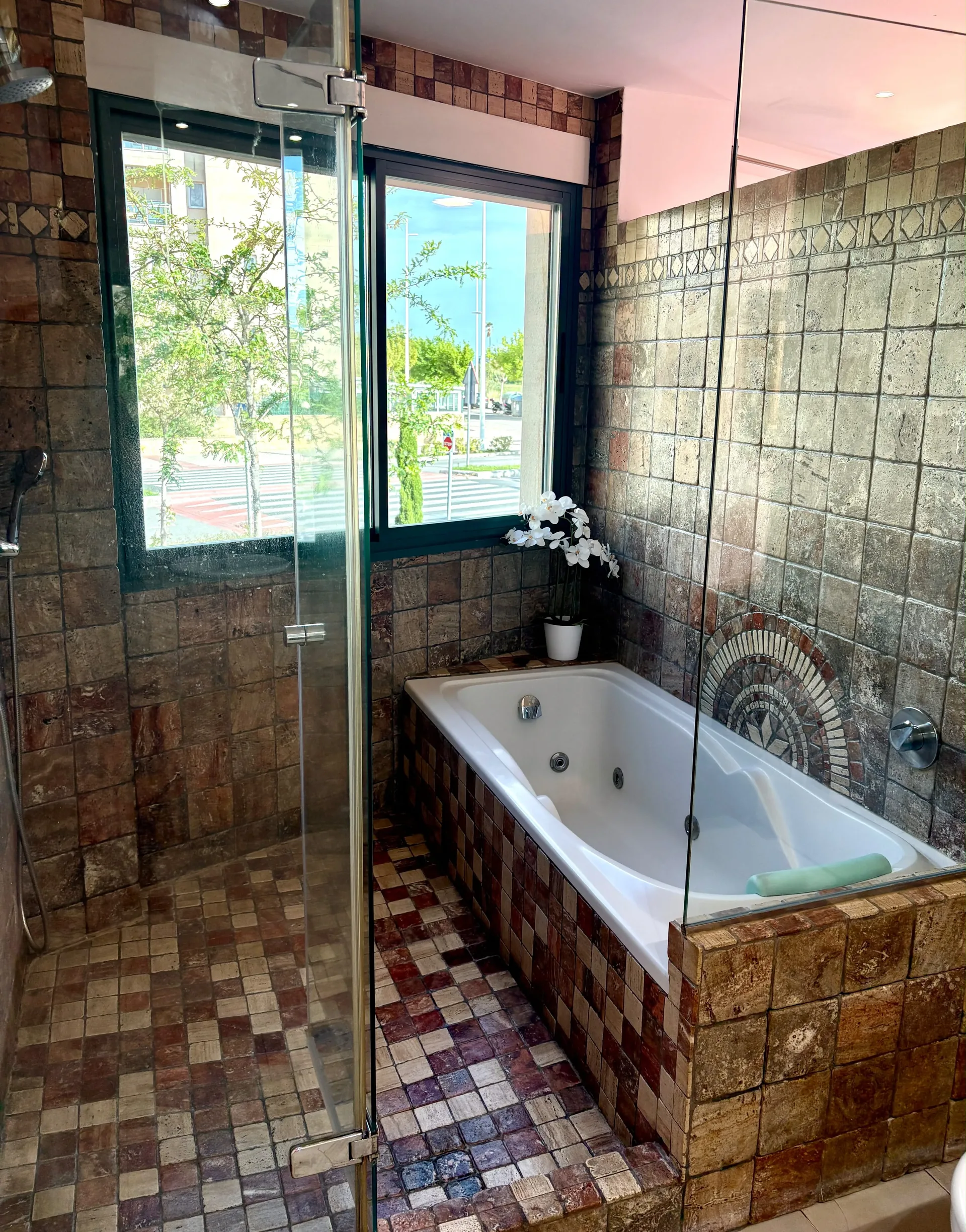 Baño con bañera de hidromasaje y ducha de cristal, azulejos de mosaico marrón y ventana al jardín — Chalet Treviso Gandía